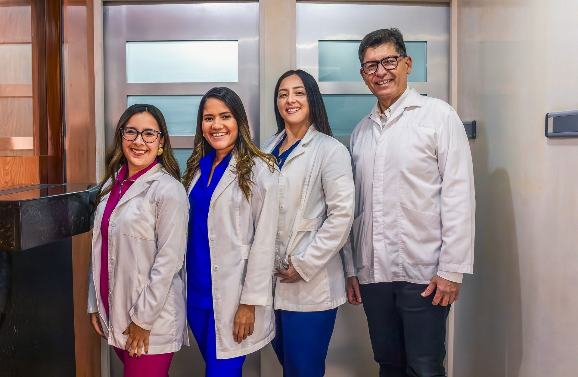 Group of four medical professionals—three women and one man—standing in a clinic, smiling at the camera, wearing scrubs and lab coats, conveying teamwork and patient care