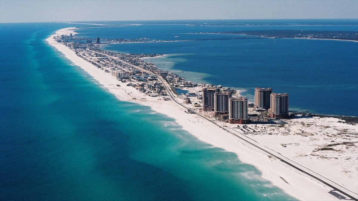 Aerial view of a long sandy barrier island with turquoise waters, high-rise buildings, and a causeway connecting to the mainland